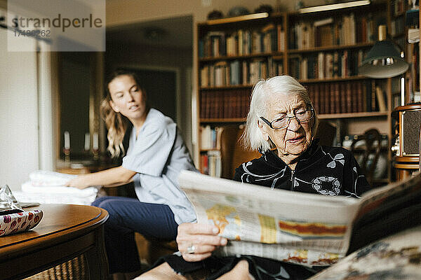 Ältere Frau liest Zeitung  während eine Betreuerin sie im Wohnzimmer beobachtet
