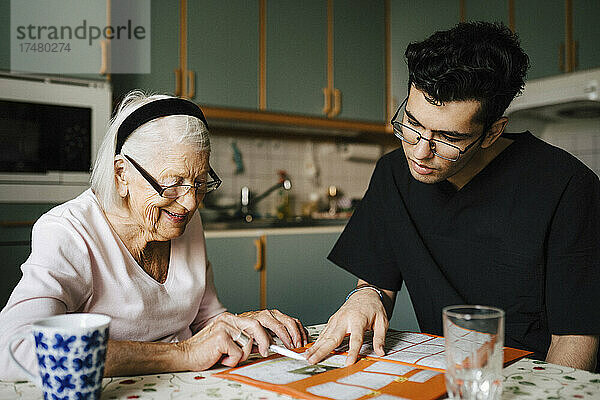 Junger Mann aus dem Gesundheitswesen spielt Kreuzworträtsel mit einer älteren Frau in der Küche