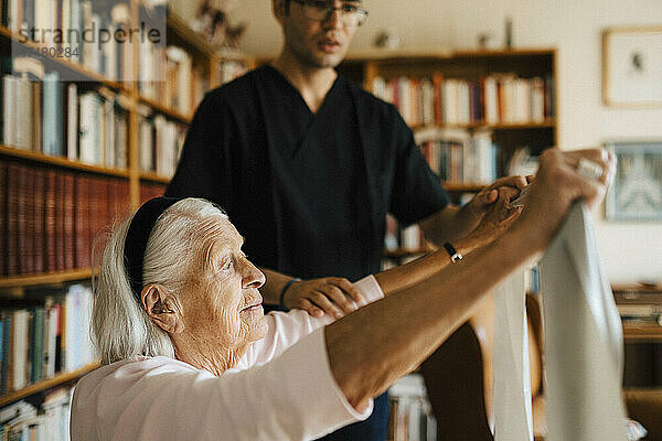 Ältere Frau macht Übungen mit einem Widerstandsband  während ein Pfleger ihr zu Hause hilft