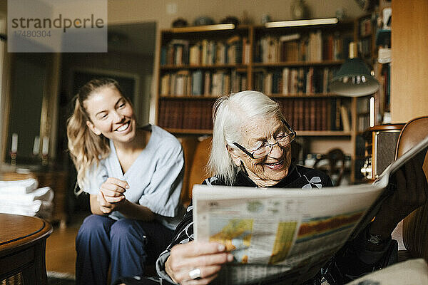 Lächelnde ältere Frau  die Zeitung liest  während im Hintergrund eine glückliche Betreuerin zu Hause ist