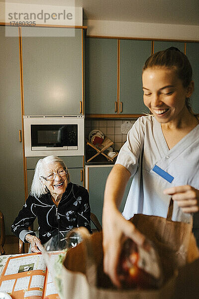 Glückliche ältere Frau  die eine Mitarbeiterin des Gesundheitswesens betrachtet  die in der Küche Lebensmittel aus der Tüte nimmt