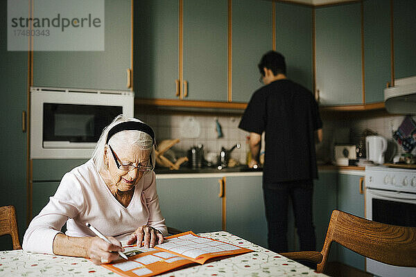 Ältere Frau löst ein Kreuzworträtsel in einem Buch  während ein Pfleger in der Küche arbeitet