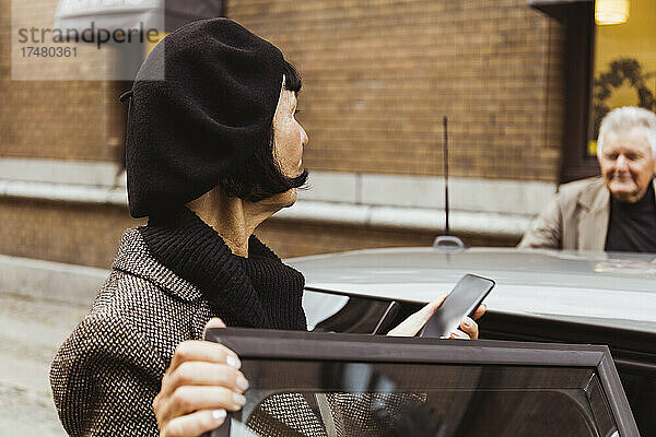 Ältere Frau mit Smartphone schaut Mann beim Einsteigen ins Auto in der Stadt an