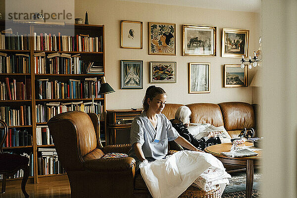 Eine Krankenschwester faltet eine Decke  während eine ältere Frau im Wohnzimmer Zeitung liest