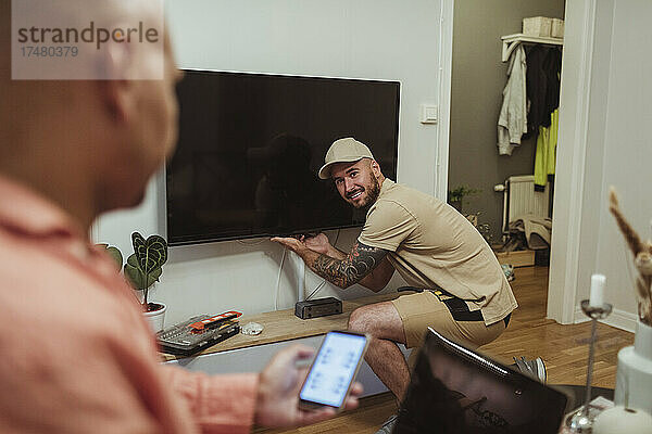 Lächelnder männlicher Techniker schaut auf einen reifen Mann  der ein Smartphone im Wohnzimmer benutzt