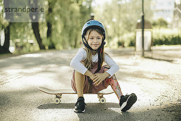 Lächelndes Mädchen mit Beinprothese  das auf einem Skateboard sitzt und nachdenkt