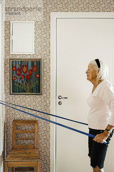 Seitenansicht einer älteren Frau  die an einem Widerstandsband zieht  während sie zu Hause trainiert