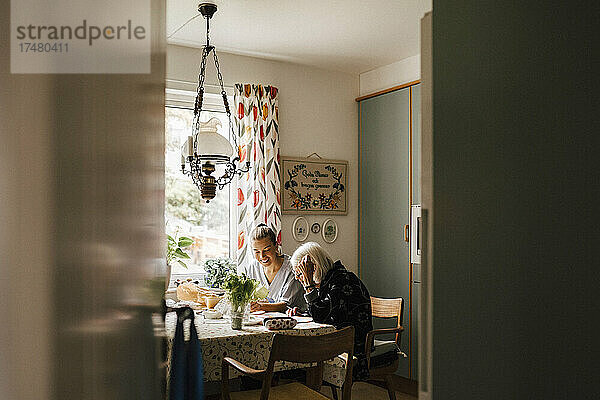 Ältere Frau  die in der Küche zu Hause bei einer weiblichen Pflegekraft sitzt und Zeitung liest