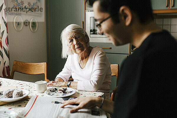 Junger Krankenpfleger liest Zeitung  während eine ältere Frau ihn in der Küche anschaut