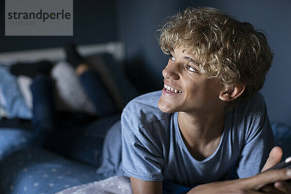 Blonder Junge lächelnd auf dem Bett liegend im Schlafzimmer zu Hause