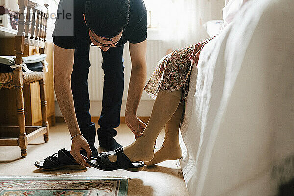 Junge männliche Pflegekraft hilft einer älteren Frau  im Schlafzimmer eine Sandale zu tragen