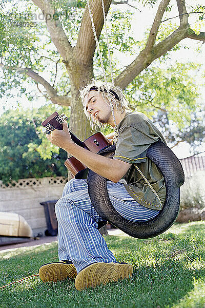 Gitarre spielender Mann  der auf einer Reifenschaukel in einem Garten sitzt und singt.