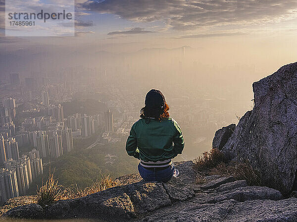 Frau sitzt auf einem Felsen in den Hügeln über Hongkong und blickt auf die Stadt hinunter.