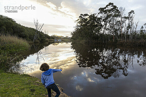 Ein Junge  der in der Abenddämmerung an einem Fluss steht  der Himmel spiegelt sich im flachen  ruhigen Wasser