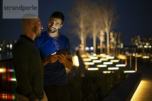 Glückliche Männer  die ein digitales Tablet in einem beleuchteten Stadtpark bei Nacht benutzen