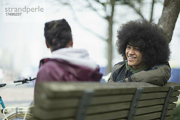 Glücklicher junger Mann mit Afro im Gespräch mit einem Freund auf einer Parkbank