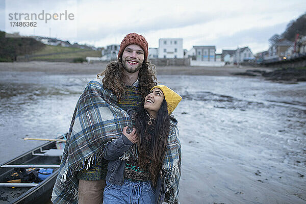Portrait glückliches junges Paar eingewickelt in eine Decke am Winterstrand