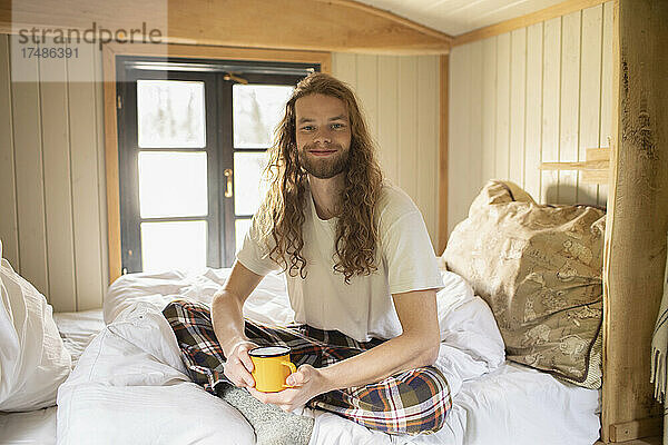 Porträt glücklicher junger Mann im Pyjama  der im Bett Kaffee trinkt