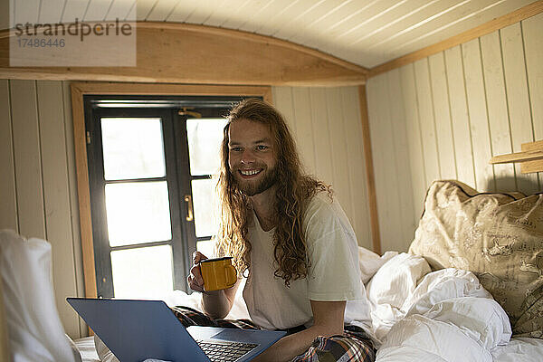 Portrait glücklicher junger Mann mit Kaffee und Laptop im Bett
