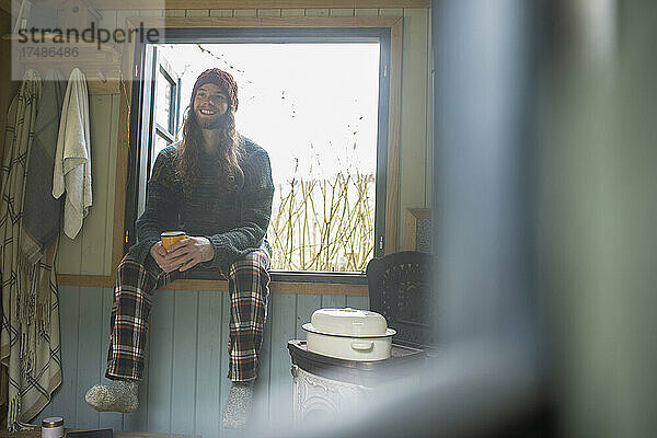 Fröhlicher junger Mann im Schlafanzug genießt Kaffee am Kabinenfenster