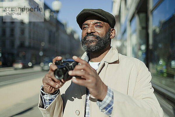 Männlicher Tourist mit Bart benutzt eine Digitalkamera auf einem sonnigen Bürgersteig