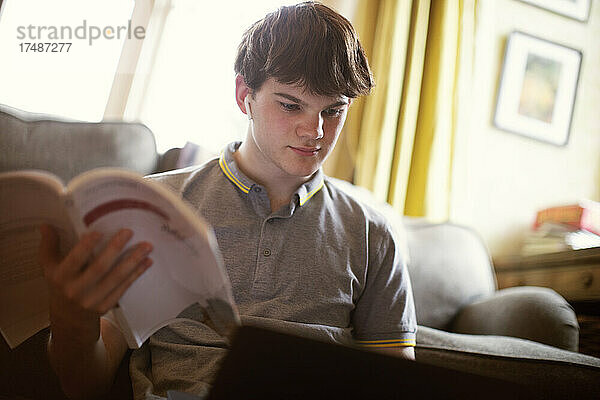 Konzentrierter Jugendlicher mit Lehrbuch und Laptop beim Lernen zu Hause
