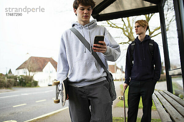 Teenager mit Skateboard und Smartphone an der Bushaltestelle