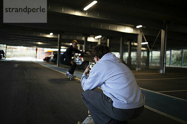 Jugendlicher mit Fotohandy filmt Freund beim Skateboardfahren auf einem Parkplatz