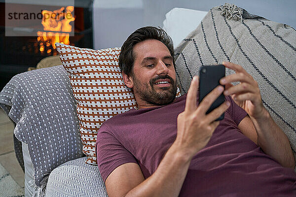 Mann mit Smartphone entspannt auf Wohnzimmersofa am Kamin