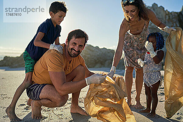 Porträt einer glücklichen Familie  die am sonnigen Strand Müll aufsammelt