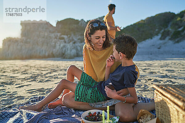 Glückliche Mutter und Sohn essen auf einer Picknickdecke am sonnigen Strand