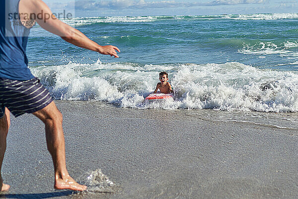 Vater beobachtet glücklichen Sohn beim Bodyboarden in der sonnigen Meeresbrandung