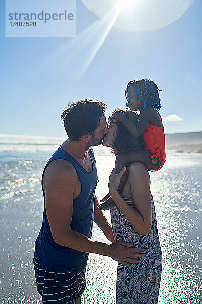 Zärtliches Paar mit Tochter küsst sich am sonnigen Meeresstrand
