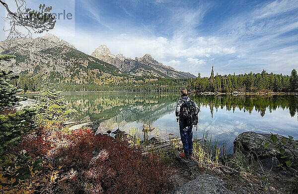 Junger Mann steht an einem See  Spiegelung im Taggart Lake  Blick auf den Teton Range Gebirgszug  Gipfel Grand Teton und Teewinot Mountain  Grand Teton National Park  Wyoming  USA  Nordamerika