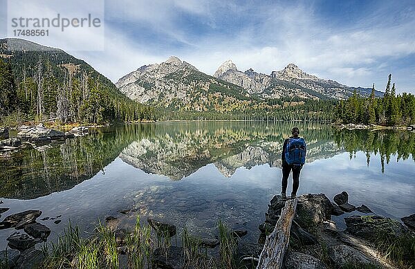 Junge Frau steht an einem See  Spiegelung im Taggart Lake  Blick auf den Teton Range Gebirgszug  Gipfel Grand Teton und Teewinot Mountain  Grand Teton National Park  Wyoming  USA  Nordamerika