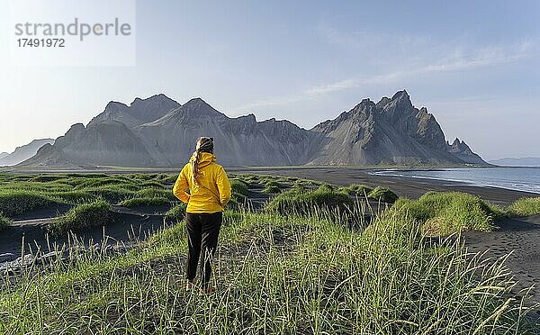 Junge Frau mit Regenjacke wandert  Schwarzer Lavastrand  Sandstrand  Dünen mit trockenem Gras  Berge Klifatindur  Eystrahorn und Kambhorn  Landzunge Stokksnes  Bergmassiv Klifatindur  Austurland  Ostisland  Island  Europa