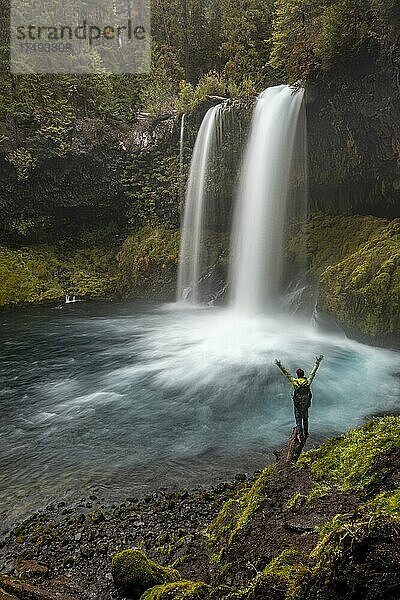Junger Mann streckt die Arme in die Luft  Wasserfall in dichter Vegetation  Koosah Falls  Oregon  USA  Nordamerika