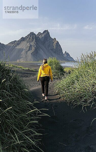 Junge Frau mit Regenjacke wandert  Schwarzer Lavastrand  Sandstrand  Dünen mit trockenem Gras  Landzunge Stokksnes  Bergmassiv Klifatindur  Austurland  Ostisland  Island  Europa