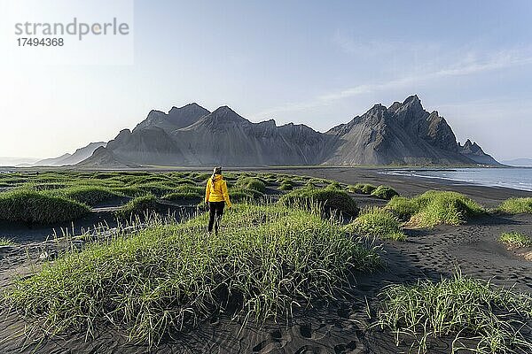 Junge Frau mit Regenjacke wandert  Schwarzer Lavastrand  Sandstrand  Dünen mit trockenem Gras  Berge Klifatindur  Eystrahorn und Kambhorn  Landzunge Stokksnes  Bergmassiv Klifatindur  Austurland  Ostisland  Island  Europa