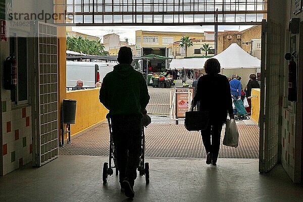 Mann mit Kinderwagen und Frau mit Einkaufstüten verlassen Markthalle  im Gegenlicht