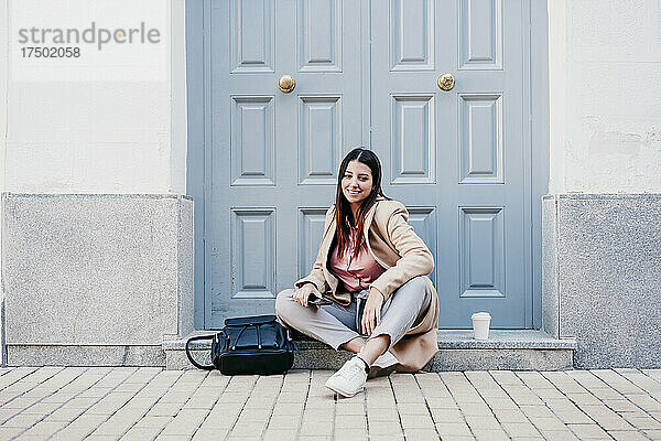 Lächelnde Geschäftsfrau sitzt mit Rucksack vor der Haustür