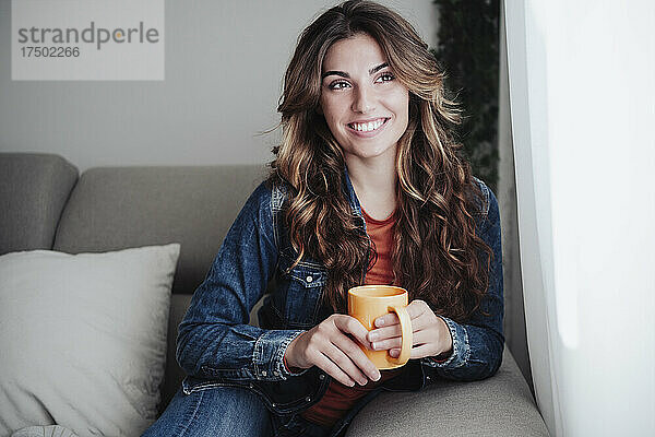 Lächelnde Frau mit Kaffeetasse sitzt auf dem Sofa am Fenster