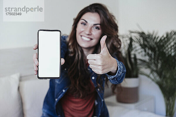 Lächelnde Frau zeigt Daumen hoch und leeren Handybildschirm im Schlafzimmer