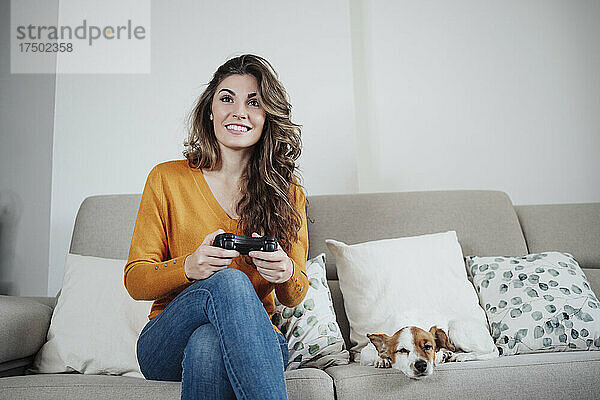 Schöne Frau spielt Videospiel und sitzt zu Hause mit Hund auf dem Sofa