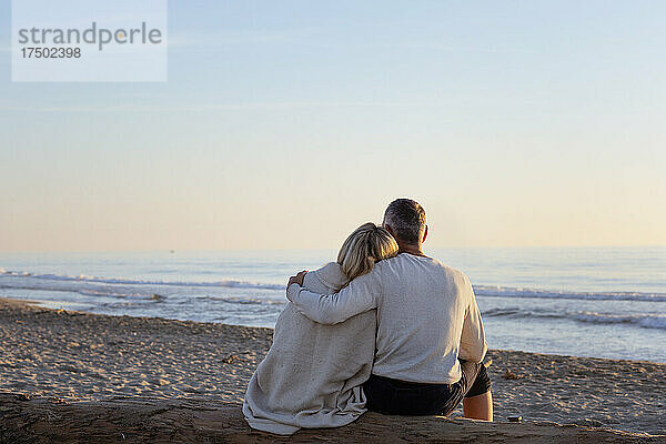 Mann mit Arm um Frau  der den Sonnenuntergang am Strand betrachtet