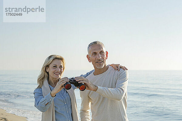 Frau mit Arm um Mann  der am Strand ein Fernglas teilt