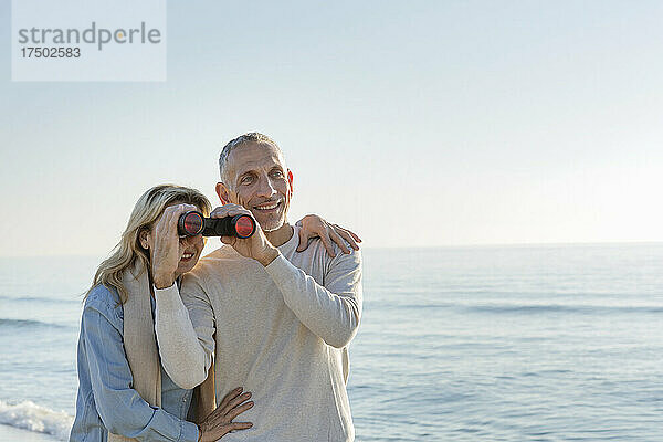 Frau blickt durch ein Fernglas  das ihr Ehemann am Strand hält