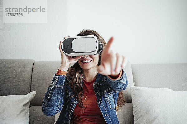 Lächelnde junge Frau mit Virtual-Reality-Headset gestikuliert auf dem Sofa