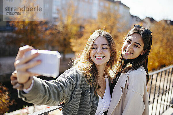 Fröhliches Teenager-Mädchen  das mit einem Freund am Geländer ein Selfie per Smartphone macht