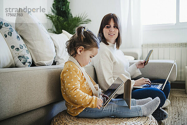 Frau betrachtet kleines Mädchen mit Tablet-PC im Wohnzimmer